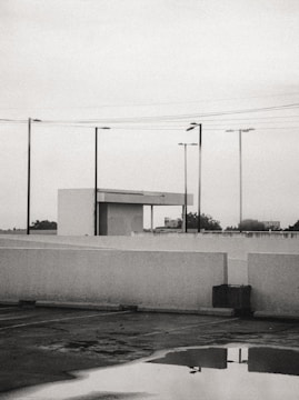A parking lot is shown with a reflective puddle on the ground, surrounded by white concrete barriers. Several streetlights and power lines intersect the overcast sky. In the background, a small building structure with a flat roof is visible, while trees and a billboard can be seen in the distance.