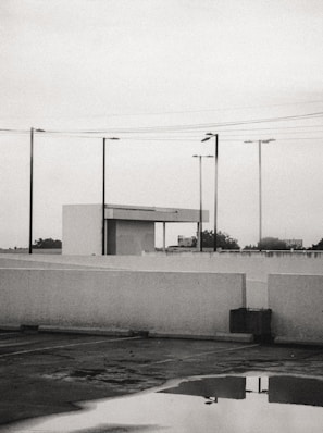 A parking lot is shown with a reflective puddle on the ground, surrounded by white concrete barriers. Several streetlights and power lines intersect the overcast sky. In the background, a small building structure with a flat roof is visible, while trees and a billboard can be seen in the distance.
