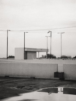 A parking lot is shown with a reflective puddle on the ground, surrounded by white concrete barriers. Several streetlights and power lines intersect the overcast sky. In the background, a small building structure with a flat roof is visible, while trees and a billboard can be seen in the distance.