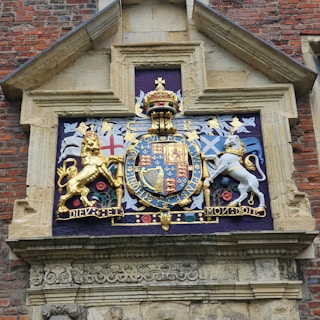 An ornate, colorful royal coat of arms is embedded in a stone wall. It features a crowned shield at the center, flanked by a golden lion on the left and a white unicorn on the right. Above the shield is a golden crown, and beneath is a motto inscribed on a ribbon. The background includes symbolic flags and decorative elements in blue, red, and gold, highlighting historical and heraldic symbols.