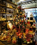 A festive store display centered around a decorated Christmas tree, adorned with ornaments such as miniature nutcrackers, candy canes, and stars. The surrounding area is filled with holiday-themed figurines, candles, and various decorative items. The shelves in the background contain a mix of antique-style items and more Christmas decorations, adding to the festive atmosphere.