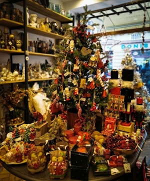 A festive store display centered around a decorated Christmas tree, adorned with ornaments such as miniature nutcrackers, candy canes, and stars. The surrounding area is filled with holiday-themed figurines, candles, and various decorative items. The shelves in the background contain a mix of antique-style items and more Christmas decorations, adding to the festive atmosphere.