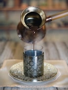An ornate metal cup is being filled with a dark liquid from an elegant, long-handled pot. The cup sits on an intricately designed saucer placed on a patterned tablecloth. The setting is elegant and suggests traditional or cultural significance.