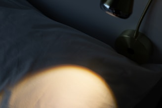 A dimly lit scene featuring a lamp casting a warm light onto a fabric surface, likely a bed or pillow. The lamp is metallic and has a modern design with a visible bulb extending from its base.