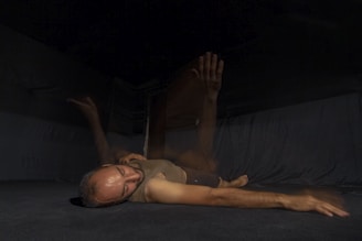 A person lying on a dark floor with a blurred motion effect, appearing to move their arm through the air. The background is dimly lit, creating a mysterious and introspective atmosphere.