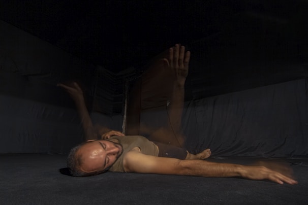 A person lying on a dark floor with a blurred motion effect, appearing to move their arm through the air. The background is dimly lit, creating a mysterious and introspective atmosphere.