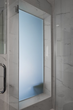 A frosted glass window set within a marble-tiled wall, featuring a chrome handle on the left. The surrounding walls have a polished marble finish with gray veining, adding to the modern aesthetic of the space.