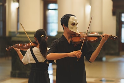 Two people are playing violins in an elegant room with large windows. One person is wearing a distinctive, theatrical mask, adding a dramatic element to the scene.