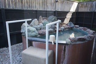 A round, elevated hot tub is surrounded by a border of natural stones. The tub is situated in a fenced backyard area with a gravel ground. White railings provide support for entering or exiting the tub. A small plastic scoop is placed on the edge of the tub.