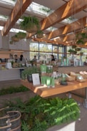 A modern store interior featuring a variety of green products displayed on wooden tables. The space is adorned with hanging plants and large windows that allow natural light to flood in, creating a warm and inviting atmosphere. The ceiling has wooden beams with skylights. Customers and staff are visible, engaged in transactions.