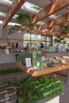 A modern store interior featuring a variety of green products displayed on wooden tables. The space is adorned with hanging plants and large windows that allow natural light to flood in, creating a warm and inviting atmosphere. The ceiling has wooden beams with skylights. Customers and staff are visible, engaged in transactions.