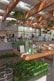 A modern store interior featuring a variety of green products displayed on wooden tables. The space is adorned with hanging plants and large windows that allow natural light to flood in, creating a warm and inviting atmosphere. The ceiling has wooden beams with skylights. Customers and staff are visible, engaged in transactions.