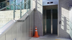 An outdoor elevator entrance made of glass surrounded by a brick wall and metal panels. A staircase with glass railing runs alongside it. A bright orange traffic cone is placed on the ground near the elevator, and shadows are cast on the wall.