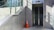 An outdoor elevator entrance made of glass surrounded by a brick wall and metal panels. A staircase with glass railing runs alongside it. A bright orange traffic cone is placed on the ground near the elevator, and shadows are cast on the wall.