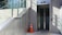 An outdoor elevator entrance made of glass surrounded by a brick wall and metal panels. A staircase with glass railing runs alongside it. A bright orange traffic cone is placed on the ground near the elevator, and shadows are cast on the wall.