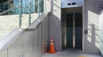 An outdoor elevator entrance made of glass surrounded by a brick wall and metal panels. A staircase with glass railing runs alongside it. A bright orange traffic cone is placed on the ground near the elevator, and shadows are cast on the wall.