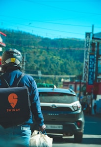 A delivery person with a Swiggy backpack is walking on a street, carrying a paper bag. The street is bustling with activity, including a car and various signage on buildings. In the background, there are hills under a clear blue sky.