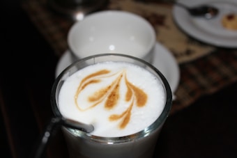 A glass of coffee with a beautifully crafted latte art design on the frothy milk surface. The background features a blurred setting with white cups and a spoon visible.