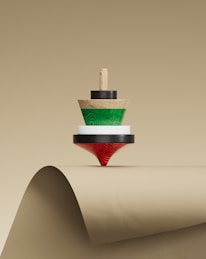 A wooden spinning top with concentric layers of different colors, including natural wood, green, white, and red, is balanced on the curved edge of a beige paper surface.