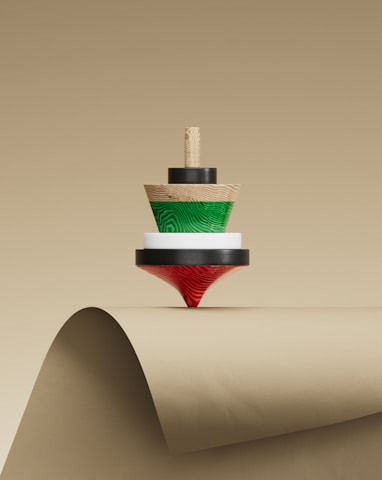 A wooden spinning top with concentric layers of different colors, including natural wood, green, white, and red, is balanced on the curved edge of a beige paper surface.