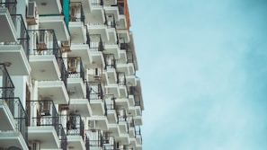 A tall apartment building with multiple balconies extends vertically, featuring white walls and black railings. Each balcony has an air conditioning unit, and the sky in the background is clear and blue.