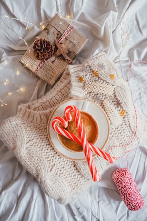 A cozy arrangement on a white textured surface features a thick, cream-colored knit sweater with a cup of coffee placed on top. Two red and white candy canes are resting on the edge of the cup. Nearby, there's a wrapped gift adorned with red and white string and a pine cone. Soft string lights are scattered around, adding a warm and festive touch.
