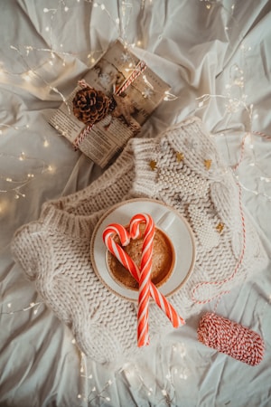 A cozy winter scene with a white knitted sweater, a steaming cup of coffee topped with two candy canes, and a wrapped gift adorned with pine cones and star-shaped decorations. Soft fairy lights are scattered throughout, creating a warm, festive atmosphere.