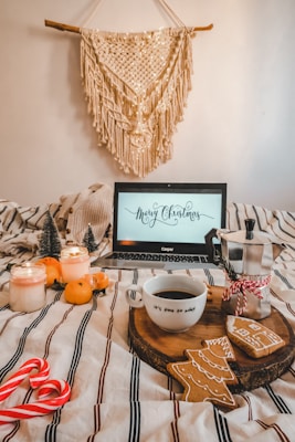 A cozy Christmas scene with a laptop displaying a 'Merry Christmas' message. The setting includes a cup of coffee on a wooden tray with Christmas-themed cookies, surrounded by festive ornaments like small decorative trees, candles, and candy canes. An orange plaid blanket covers the bed, and a macram&eacute; wall hanging adds a warm touch.
