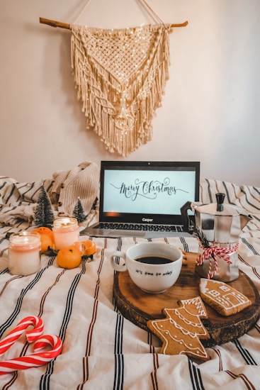 A cozy Christmas scene with a laptop displaying a 'Merry Christmas' message. The setting includes a cup of coffee on a wooden tray with Christmas-themed cookies, surrounded by festive ornaments like small decorative trees, candles, and candy canes. An orange plaid blanket covers the bed, and a macram&eacute; wall hanging adds a warm touch.