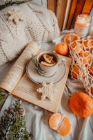 A cozy arrangement featuring an open book with a cup of tea or coffee resting on it, garnished with lemon slices and a sprig of rosemary. A knitted pillow and several books are nearby, along with a lit candle labeled 'Be still.' Fresh oranges and peeled orange segments are scattered, some in a net bag. A decorative snowflake cookie adds a festive touch.