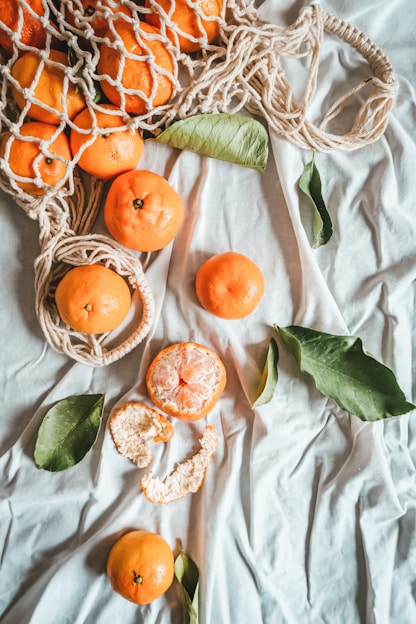 A collection of bright orange tangerines is scattered across a light blue fabric surface. Some of the fruits are enclosed in a netted, rope bag, while others are placed individually. One tangerine is partially peeled, showing its juicy segments. Green leaves are interspersed among the tangerines, providing a natural contrast.