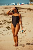 A woman in a black swimsuit stands on a sandy beach with seaweed scattered around. She has a tattoo on her arm and is holding a pair of flip-flops in one hand while shielding her eyes from the sun with the other. In the background, beach umbrellas and chairs are visible.