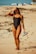 A woman in a black swimsuit stands on a sandy beach with seaweed scattered around. She has a tattoo on her arm and is holding a pair of flip-flops in one hand while shielding her eyes from the sun with the other. In the background, beach umbrellas and chairs are visible.