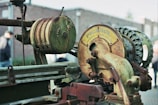 A close-up of an old, rusted mechanical device with visible gears and a cylindrical component. The machinery includes a circular part with the brand 'Picanol' and the text 'Ypres Belgium'. The background is blurred, showing a brick wall and a person wearing a hat.