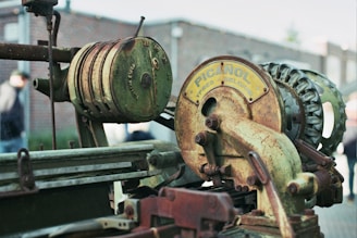 A close-up of an old, rusted mechanical device with visible gears and a cylindrical component. The machinery includes a circular part with the brand 'Picanol' and the text 'Ypres Belgium'. The background is blurred, showing a brick wall and a person wearing a hat.