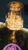 A glass pendant light fixture contains a tangled cluster of small, glowing string lights that emit a warm, golden glow. The background is blurred, providing a contrast to the detailed texture of the wires and lights within the glass.