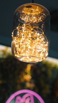 A glass pendant light fixture contains a tangled cluster of small, glowing string lights that emit a warm, golden glow. The background is blurred, providing a contrast to the detailed texture of the wires and lights within the glass.