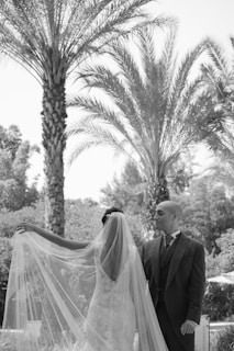 A bride and groom are standing outdoors, surrounded by tall palm trees and lush greenery. The bride is wearing a long, veil-draped gown, and the groom is dressed in a suit. The scene is peaceful and elegant, captured in black and white.