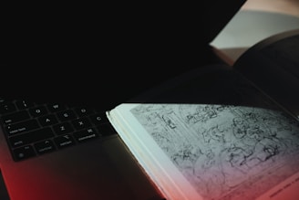 An open book with a detailed sketch lies on top of a laptop keyboard. The light focuses on the left page of the book, revealing intricate drawings, while the surrounding area is shaded. The keyboard displays keys like Control, Option, and Command.