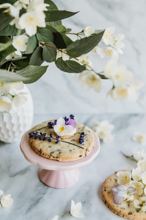 A rustic cookie garnished with purple and white flowers sits on a pastel pink pedestal, surrounded by white blossoms and green leaves on a marble surface. Another similar cookie rests nearby, adorned with delicate floral arrangements.