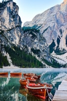 Wooden boats are docked on a serene lake surrounded by towering rocky cliffs and lush green pine trees. The water reflects the landscape and the boats, creating a mirror-like effect. The scene is peaceful, with the early morning or late afternoon light casting soft shadows.