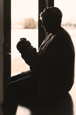 A silhouette of a person holding a baby, sitting by a window with soft light streaming through. The gentle posture conveys care and tenderness.