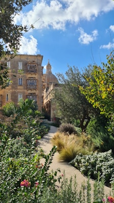 A charming garden landscape with a variety of lush green plants and colorful flowers along a winding path. In the background, an old stone building with ornate architecture and a dome is partially visible, set against a bright blue sky with scattered clouds.