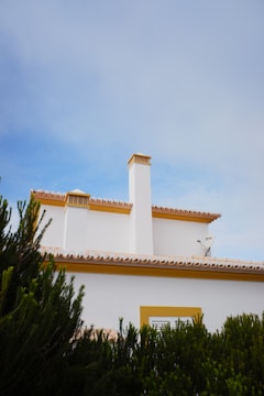 A white residential building with a yellow trim featuring multiple chimneys sits against a clear blue sky. Green foliage frames the lower part of the building and a satellite dish is attached to the side.