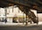 An urban street corner featuring an old, elevated train track structure made of metal, with worn and rusty beams. Below the tracks, there are staircases leading up. The storefronts below include signs for 'High Ridge News & Cards', 'Van Cortlandt Deli & Grocery', and an advertisement for Atlantic City Buses. The shops have colorful awnings and display items in their windows. A few people are seen walking or standing near the shops. The area appears to be in a city with heavy infrastructure. Traffic lights are visible hanging from the elevated structure.