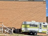 A vintage trailer with a green and silver exterior is parked near a large brick wall. There are old wooden stairs and stacked boards beside the trailer, suggesting a somewhat neglected or industrial setting. The sky is clear and blue, and there are electrical wires overhead.