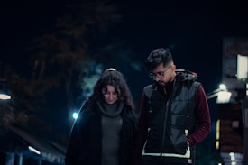 A man and a woman are walking together at night. They both wear dark, warm clothing. The setting is illuminated by streetlights, and trees can be seen blurred in the background. The couple appears to be engaged in a quiet moment or conversation.