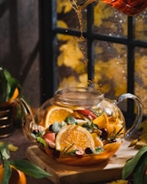 A glass teapot filled with assorted fruits such as orange slices, apples, and cranberries is placed on a wooden board. Fresh mint leaves and sprigs of rosemary add a touch of green. Tea is being poured into the teapot, and steam is visible. The background features a window with autumn leaves visible outside, casting a warm, cozy glow.