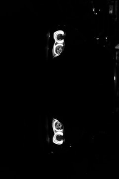 Bright, circular car headlights shine in stark contrast to the surrounding darkness. The image captures an intense focus on the twin lights, creating a symmetrical and almost mirror-like reflection.