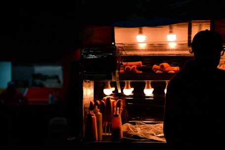A dimly lit street food stall featuring churros under warm, glowing lights. The silhouette of a person is visible next to the stall, creating a contrast with the illuminated food items. Bottles of condiments are also present on the counter.